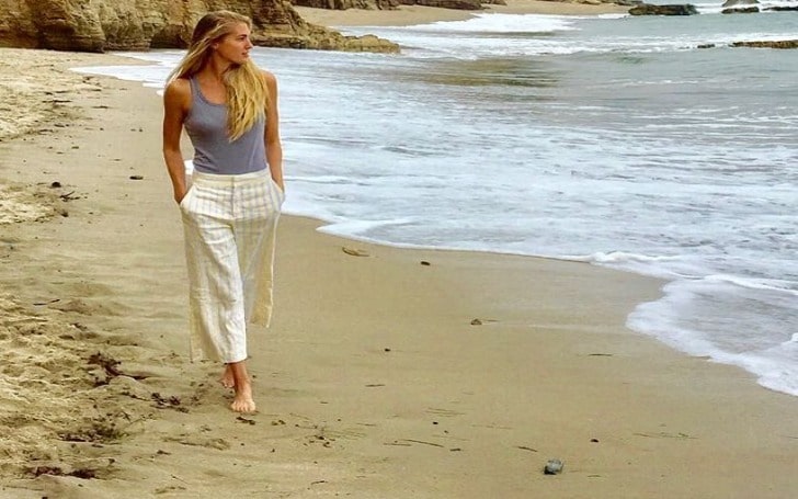 Candace Kroslak in a light purple sleeveless and white pants walking along the seashore.